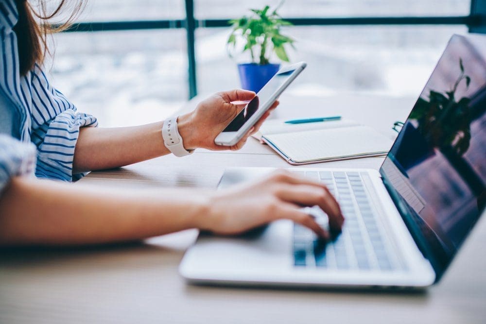 woman wearing a white watch holding a smart phone with one hand and on a laptop with the other hand.