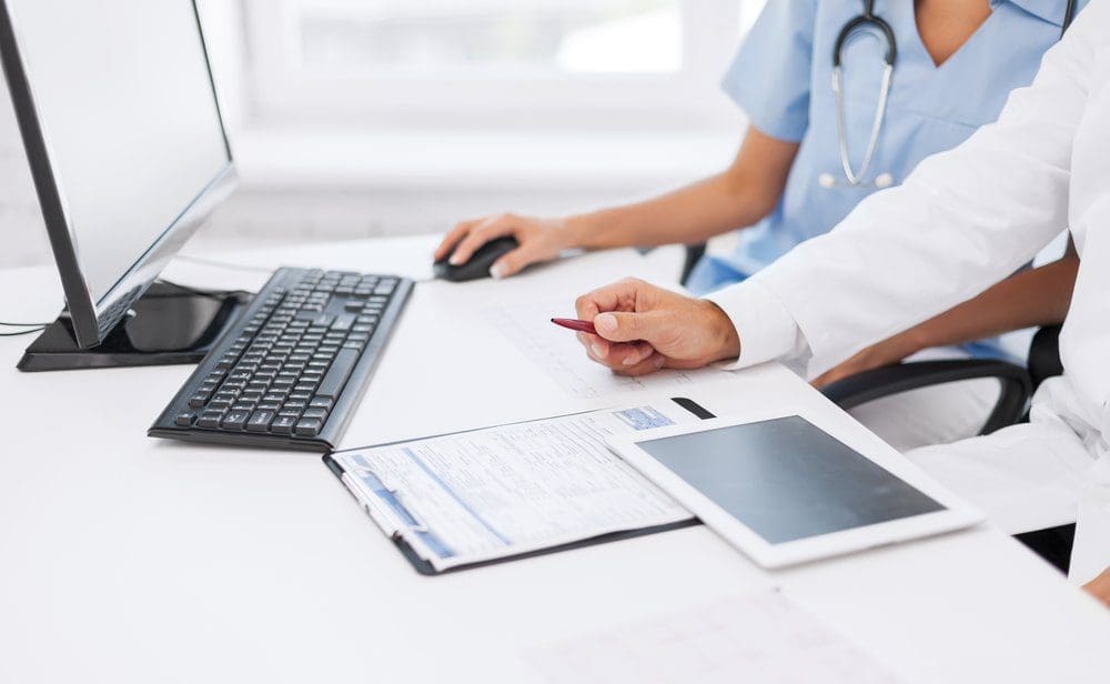 Two doctors examine medical records on a computer while sitting at a white desk and have a tablet.