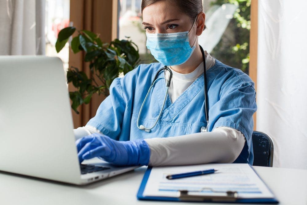 Female doctor checking up on patient records on a laptop with a clipboard and pen next to her on the table.